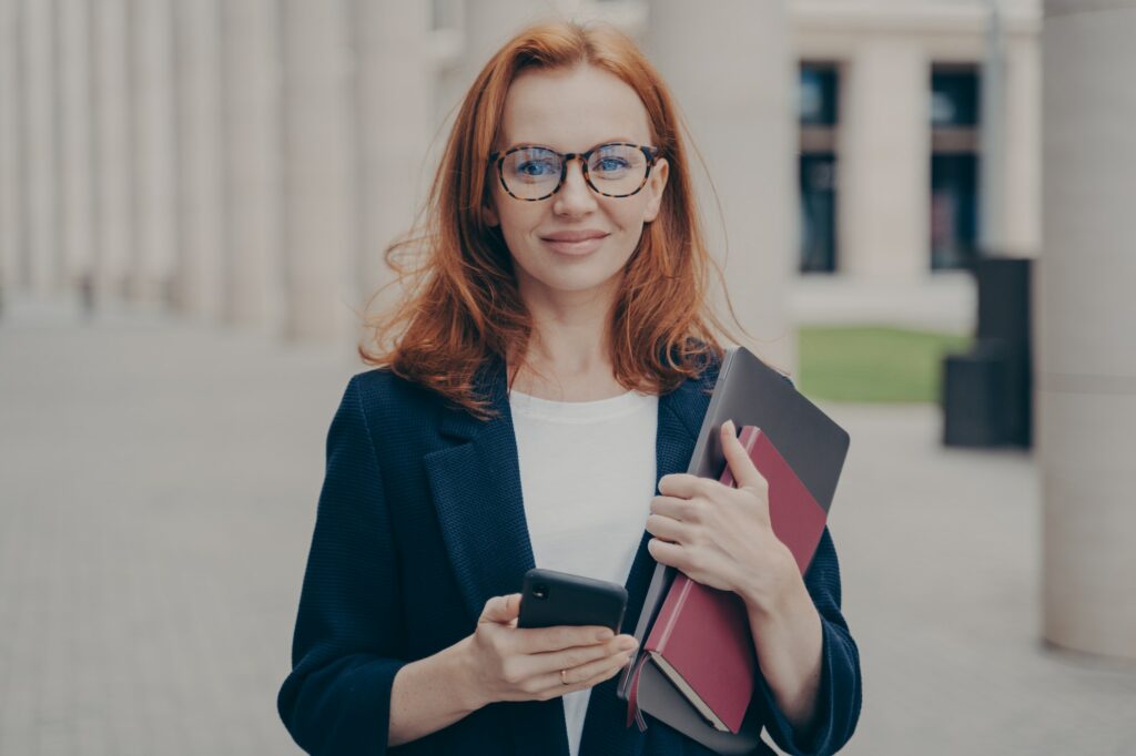 confident beautiful red haired female business consultant holding modern smartphone and laptop